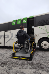 Keleivis su ribotu judumu, sėdintis neįgaliojo vežimėlyje, keliamas specialiu keltuvu į tarpmiestinį TOKS autobusą.